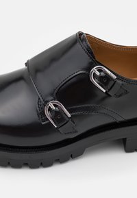 Chaussure en cuir noir avec deux boucles métalliques, texture lisse, forme arrondie et semelle en caoutchouc noir épaisse avec motif de crampons.