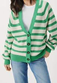 Cardigan a righe verde e bianche con scollatura a V profonda, grandi bottoni e polsini a coste, realizzato in tessuto lavorato a maglia, abbinato a una t-shirt bianca.