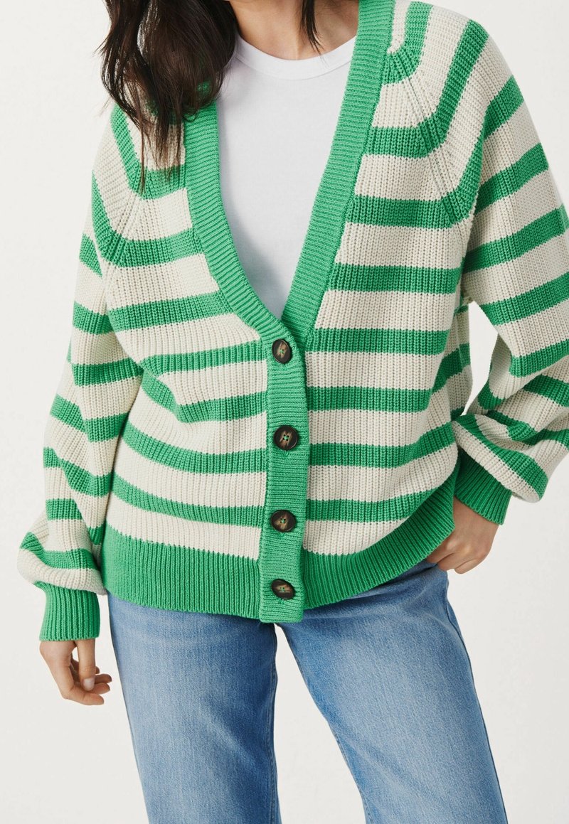 Cardigan a righe verde e bianche con scollatura a V profonda, grandi bottoni e polsini a coste, realizzato in tessuto lavorato a maglia, abbinato a una t-shirt bianca.