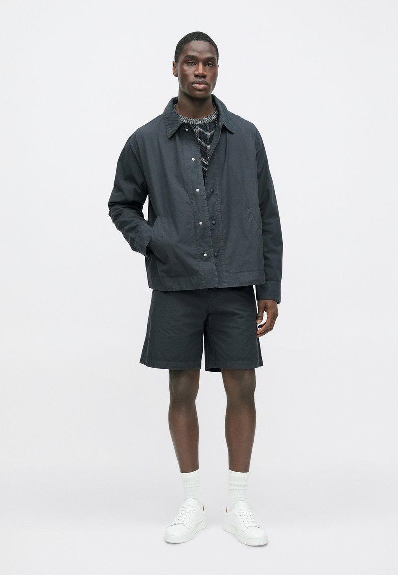 Black lightweight jacket, collared with snap buttons, worn over a patterned top, paired with black shorts and white sneakers with socks.