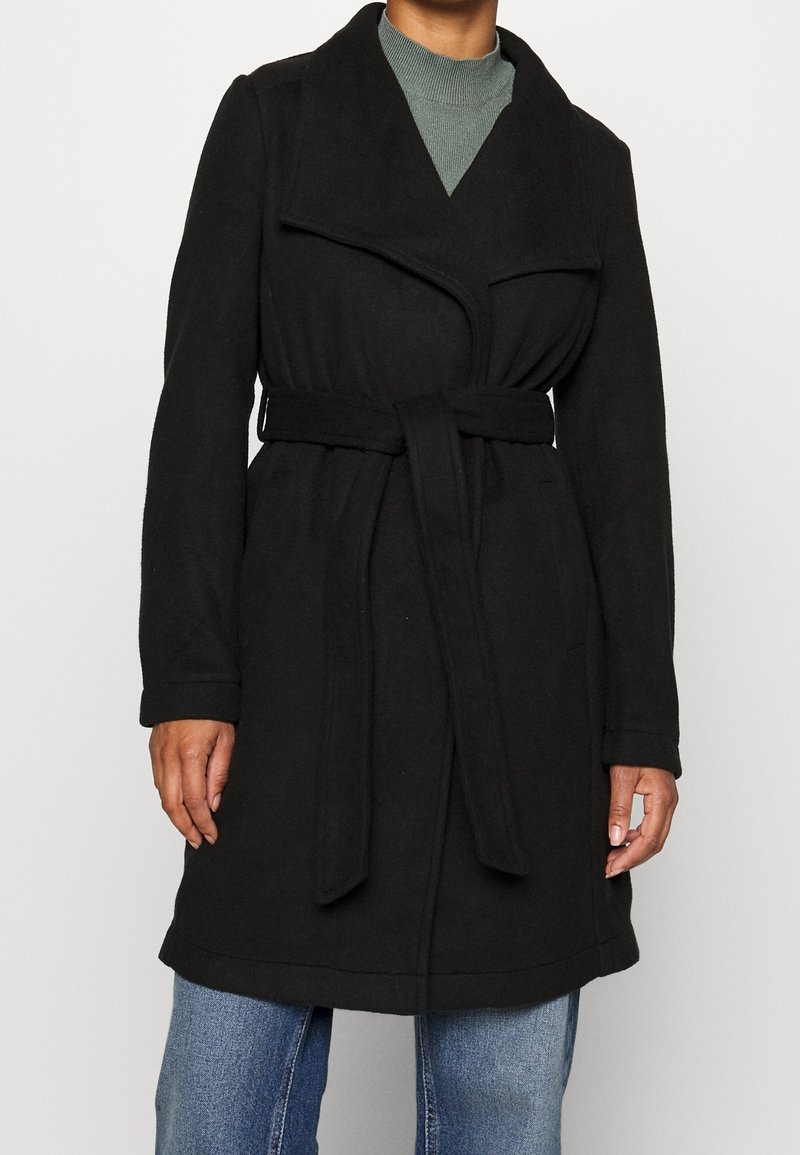 Personne portant un manteau noir ceinturé par-dessus un pull à col roulé vert et un jean bleu, debout devant un fond blanc uni.