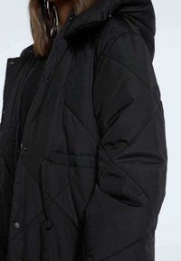 Czarna pikowana kurtka z kapturem, zatrzaskami i bocznymi kieszeniami. Materiał ma gładką tekstur�ę z geometrycznym wzorem w romby.