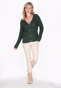 Cardigan lavorato a maglia verde bosco con scollo a V, chiusura a bottoni e un motivo intricato. Abbinato a pantaloni beige aderenti e scarpe chiare.