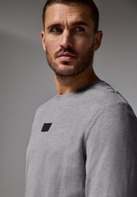 Grå långärmad skjorta i mjukt tyg med en svart etikett på bröstet. Enkel design med rund halsringning och inga mönster.