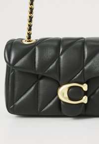 Sac à main en cuir matelassé noir avec une bandoulière en chaîne dorée et un fermoir en métal doré proéminent. La texture est lisse avec une finition rembourrée.