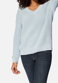 Hellblauer Strickpullover mit V-Ausschnitt, strukturiertem Muster und senkrechten Schultern. Kombiniert mit dunklen Jeans, die eine lockere Passform zeigen.