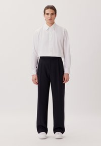 Man die recht vooruit staat, gekleed in een wit knoopshirt, zwarte wijde broek en witte sneakers, tegen een effen achtergrond.