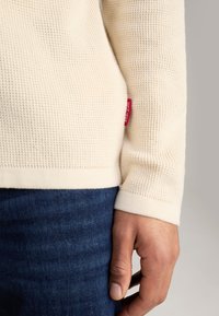 Maglione di colore crema con texture a coste, dotato di un'etichetta rossa, abbinato a jeans di denim blu scuro. Inquadratura ravvicinata del maniche e della mano.
