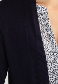 Marinblå stickad cardigan med ribbat mönster, öppen frontdesign, lager över en vit skjorta med en delikat blommig design.