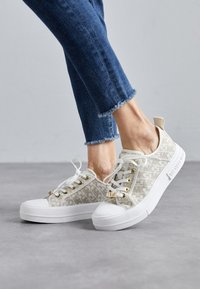 Kangasneakers i beige med logotryck, vit gummitå, vit sula, guldöglor och vita snören. Bärs med slitna blå jeans.