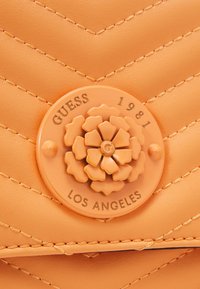 Pokrowiec w kolorze brzoskwiniowym, pikowany, z ekoskóry z motywem chevron, z pomarańczowym zapięciem w kształcie kwiatu z wytłoczonymi detalami "GUESS", "1981" i "LOS ANGELES".