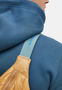 Rugzak met een gewatteerd gouden gedeelte, lichtblauwe schouderband met het label "Peak Performance," gedragen over een blauwe sweatshirt met een hoge nek.