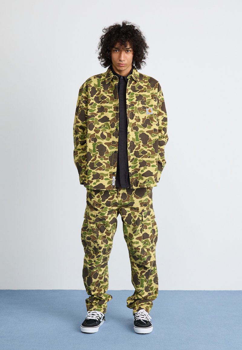 Olivenfarbene, braune und gelbe camouflierte Jacke und Hose aus einem Stoff mit strukturierter Oberfläche. Darunter ein schwarzes T-Shirt, kombiniert mit schwarzen Sneakers.