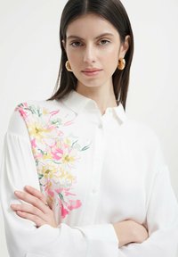 Weißes Hemd mit Knöpfen und floralem Muster in Pink, Gelb und Grün auf einer Schulter; hat lange Ärmel und einen klassischen Kragen.