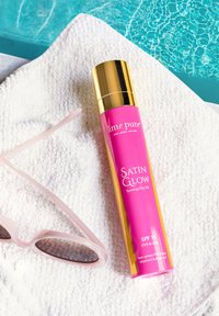 Rosa sprayflaska med guldlock märkt "Satin Glow Tanning Dry Oil," SPF 15, placerad på en vit handduk bredvid rosa solglasögon.