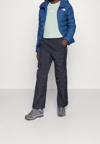 Chaqueta acolchada azul, camiseta verde claro, pantalones cargo negros y zapatos de senderismo grises. La chaqueta tiene capucha y diseño acolchado.