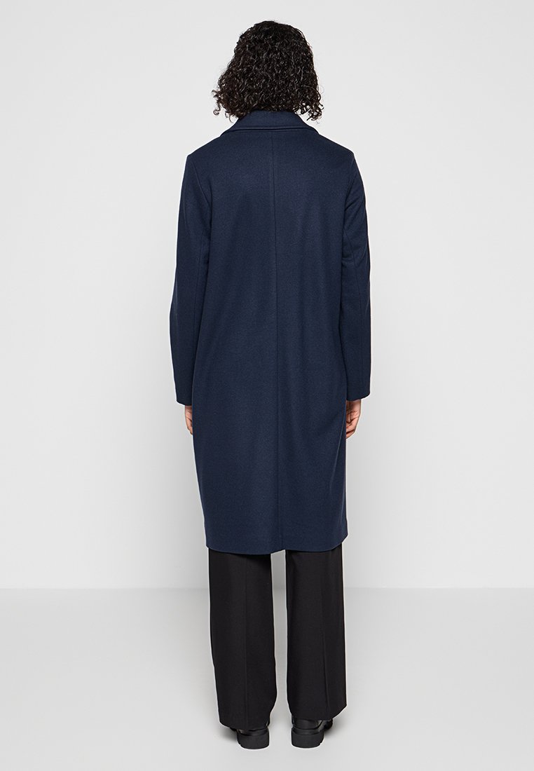 Cappotto lungo navy con taglio dritto, collo a risvolto e maniche lunghe. Realizzato in un tessuto lucido e testurizzato. Indossato sopra pantaloni neri.