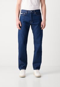 Vaqueros de mezclilla azul oscuro con un corte recto, diseño de cinco bolsillos y botón de metal. Llevados con una camiseta blanca básica y zapatillas blancas.