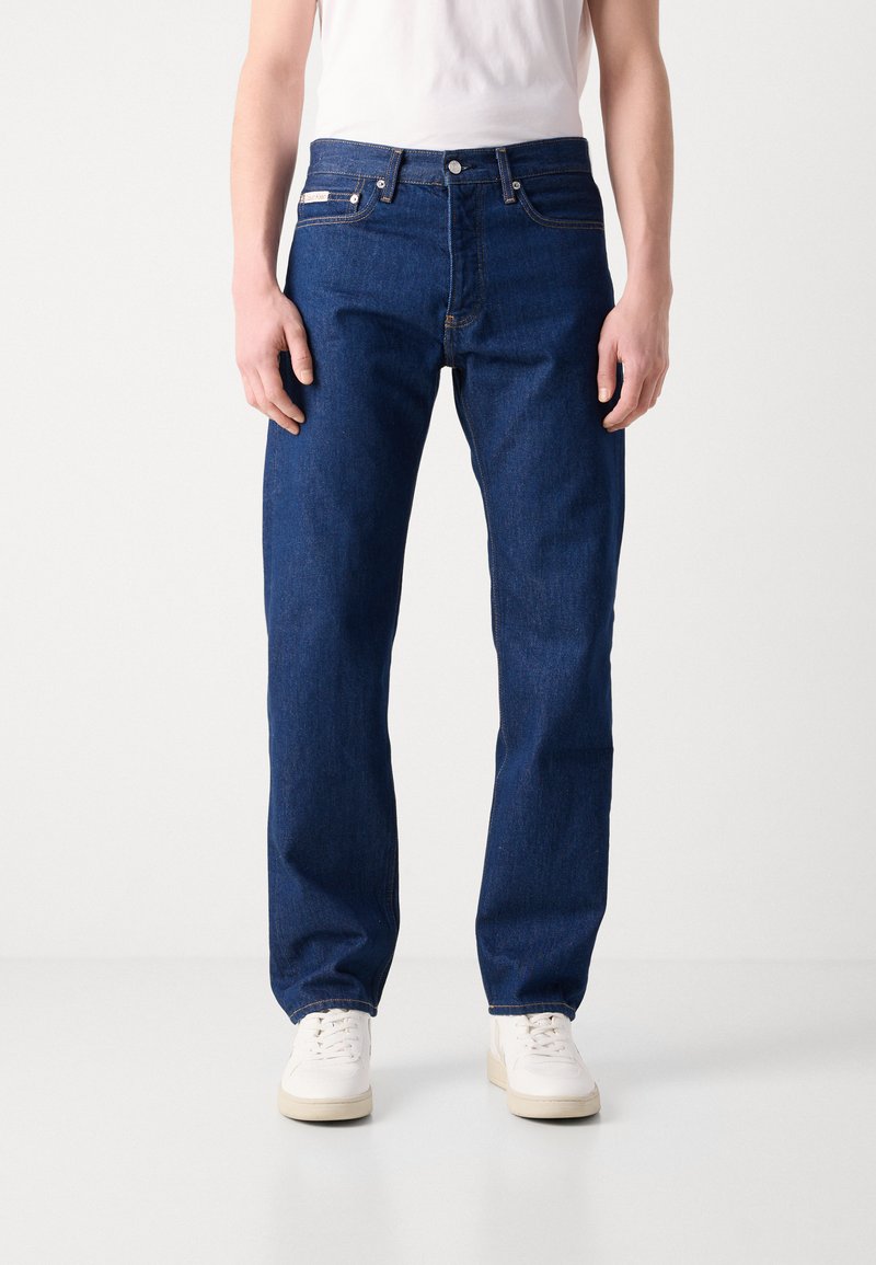 Granatowe dżinsy o prostym kroju, pięć kieszeni i metalowy guzik. Noszone z prostą białą koszulką i białymi trampkami.