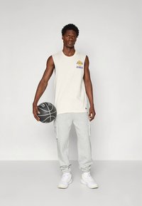 White sleeveless basketball jersey with purple and yellow logo, paired with light gray cargo sweatpants and white sneakers, holding a basketball.