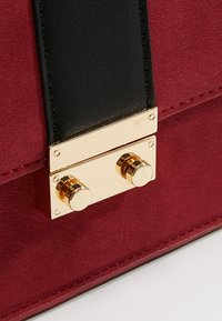 Sac en suédine fausse couleur cramoisi, doté d'une bandoulière en cuir noir et d'une quincaillerie dorée avec deux accents métalliques ronds sur la fermeture.