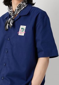 Mörkblå kortärmad skjorta med knäppning framtill, prydd med en patch-logotyp och en färgglad design, tillsammans med en svart och vit mönstrad scarfs.