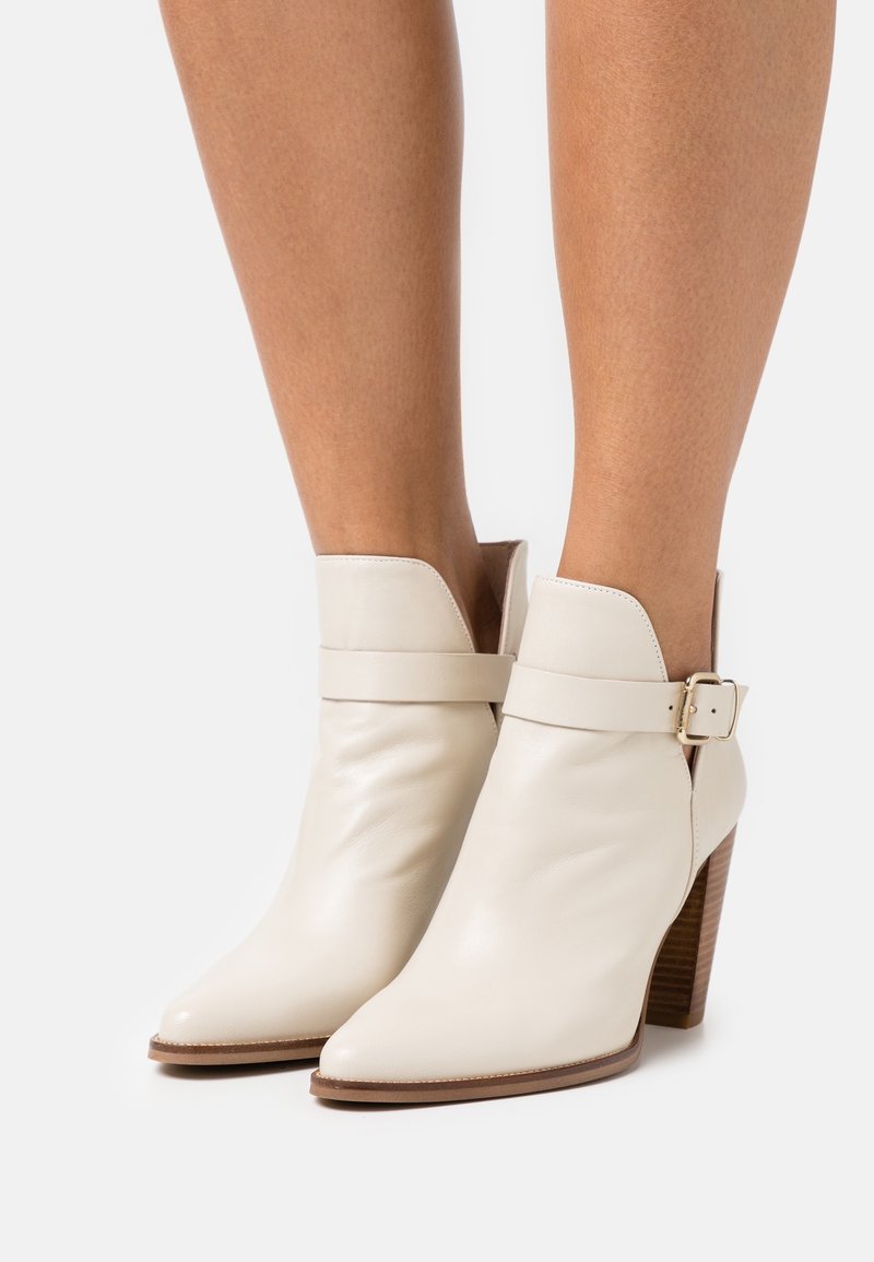 Jambes portant des bottines en cuir crème avec des bouts pointus, des talons blocs en bois et une bride latérale avec une boucle dorée.