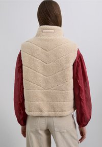 Chaleco de forro polar beige con patrón de acolchado en forma de diamante, cuello alto y parche con marca. Combinado con una blusa semitransparente roja y pantalones de pana beige.