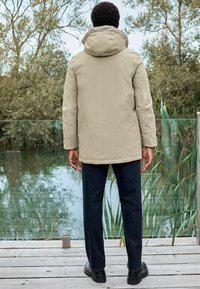 Beige Kapuzenjacke mit einem leicht lockeren Schnitt, getragen über dunklen Jeans und schwarzen Schuhen, stehend auf einer Holzplattform neben einer reflektierenden Wasseroberfläche.