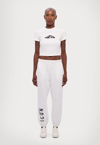 T-shirt blanc coupé avec le logo noir "ICON", associé à un pantalon de survêtement blanc ample comportant une impression noire sur le côté gauche, porté avec des chaussures blanches.