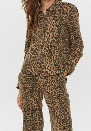 Woman wearing a matching brown and black leopard print button-up jacket and pants, standing with one hand in pocket.