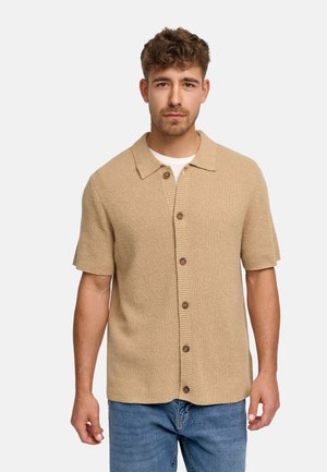 Homme aux cheveux courts et bouclés portant un cardigan beige à manches courtes boutonné par-dessus une chemise blanche et un jean bleu, debout devant un fond uni.