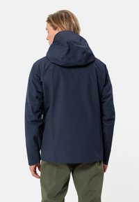 Giacca impermeabile blu navy con cappuccio, caratterizzata da una texture liscia, maniche lunghe e una vestibilità comoda, abbinata a pantaloni verde oliva.