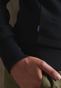 Sudadera negra con puños y dobladillo acanalados, que presenta una etiqueta azul en el costado. Enfatiza una textura suave y un diseño ajustado.