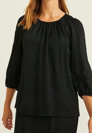 Vrouw draagt een losse zwarte blouse met driekwart ballonmouwen en lichte plooien bij de hals.