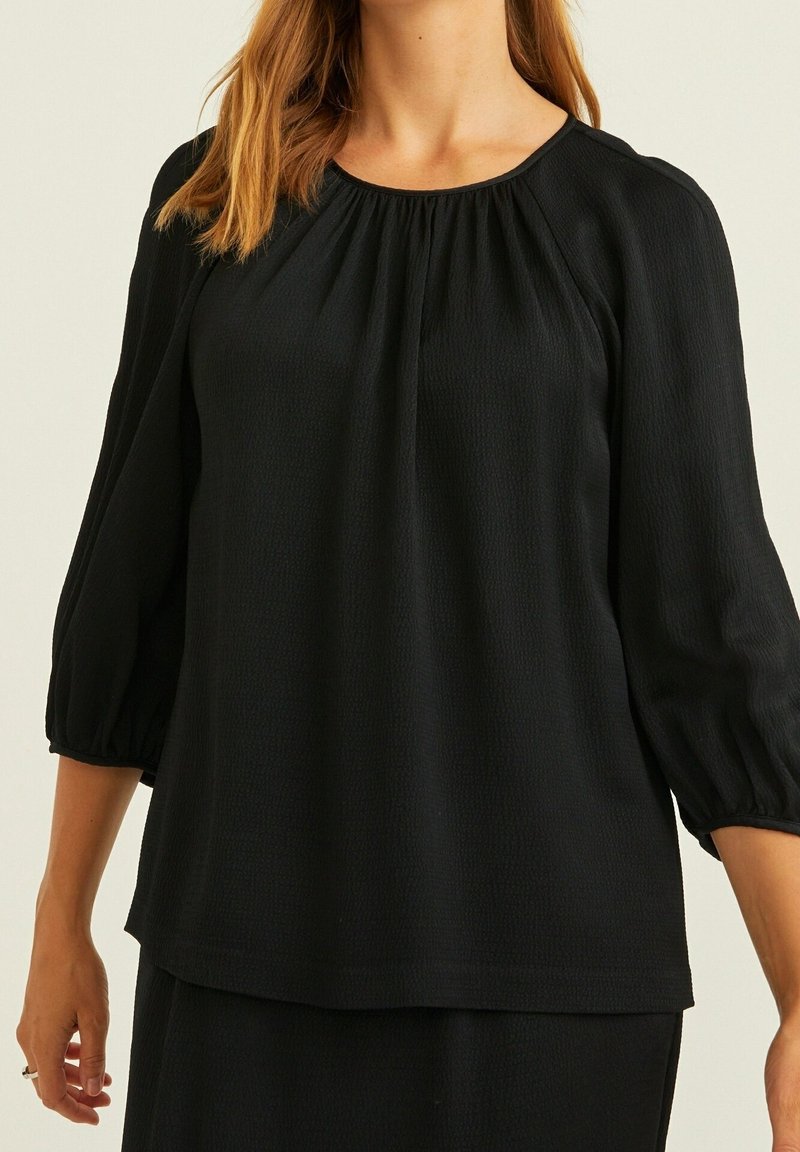 Femme portant un chemisier noir ample avec des manches ballon trois-quarts et de légers fronces au niveau de l'encolure.