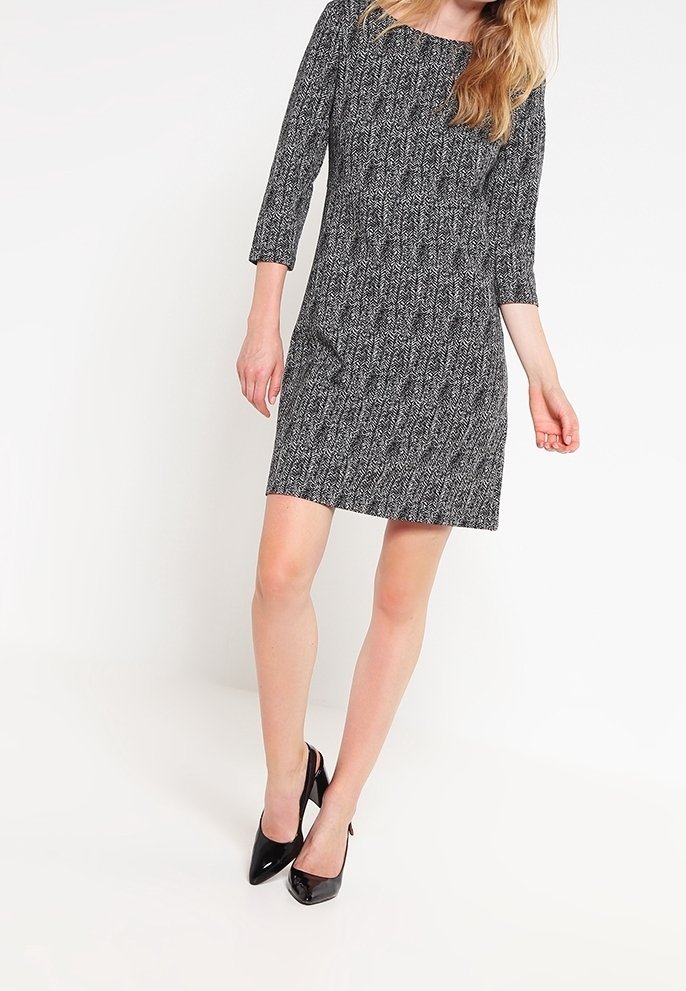 Femme portant une robe noire et blanche à motifs longueur genou avec des manches trois-quarts et des chaussures à talons noires, debout sur fond blanc.