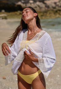 Copricostume bianco leggero con dettagli in pizzo, indossato sopra un bikini giallo. Vestibilità ampia e maniche larghe, sullo sfondo di una spiaggia sabbiosa.