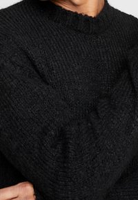 Svart stickad tröja med en texturerad mönster, rund hals och något oversized ärmar. Materialet verkar mjukt och varmt.