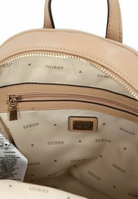 Mochila beige con exterior liso; el interior presenta un forro de tela clara con el logo "GUESS" repetido y un bolsillo con cremallera.