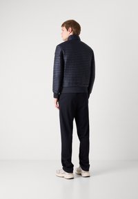 Veste rembourrée bleu marine avec des coutures horizontales, col montant et poignets côtelés, associée à un pantalon noir et des chaussures claires. Design minimaliste.