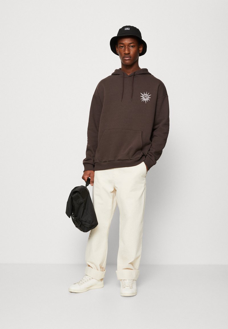 Sweat à capuche marron avec une poche avant et un motif de soleil sur la poitrine, associé à un pantalon blanc retroussé, et tenant un sac noir.