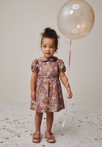 Vestido floral rosa con mangas abullonadas, cuello redondo y cintura fruncida. La niña sostiene un globo transparente con una cinta rosa; confeti brillante en el suelo.