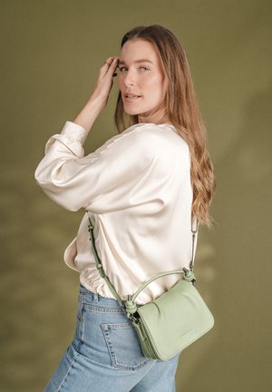 Vrouw met lang haar draagt een crèmekleurige satijnen blouse en lichtblauwe jeans, houdend een lichtgroene leren crossbodytas tegen een olijfgroene achtergrond.