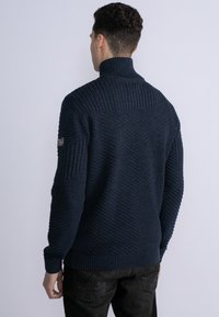 Mörkblå stickad tröja med hög krage, med diagonalt ribbade mönster och texturerad kropp, ribbade ärmar och krage.