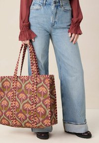 Grand sac fourre-tout à motif avec un fond marron, design floral en rose et vert, doté de poignées rayées. Associé à un jean ample bleu clair.