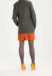 Grijze wollen jas met schouderepauletten, oranje geplooide rok, grijze panty en oranje hoge hakken, gezien van achteren.