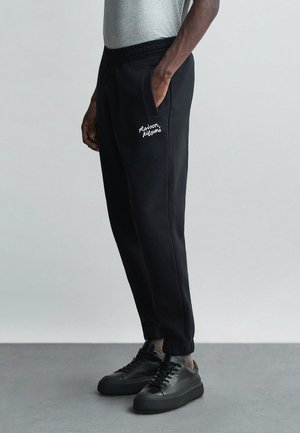 Czarne dresowe spodnie z bocznymi kieszeniami, elastycznym pasem i białym haftowanym logo "Maison Kitsuné". Noszone z czarnymi sneakersami i szarym topem.