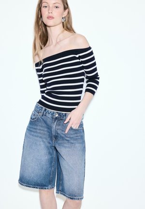 Młoda kobieta w czarno-białej bluzce w paski z odkrytymi ramionami oraz spranych, dżinsowych szortach do kolan stojąca na tle białego tła.