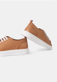 Barna bőr sportcipő alacsony profilú, fehér gumitalppal és fehér fűzőkkel. Hátoldali fül a könnyű felvétel érdekében. Simaság és minimalista design jellemzi.